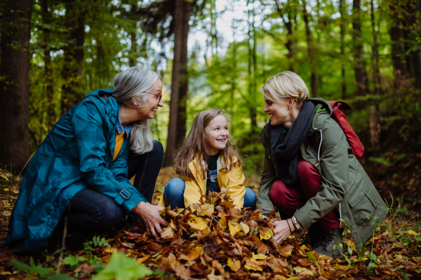 A happy little girl with mother and grandmother having fun with leaves during autumn walk in forest