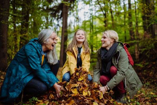 A happy little girl with mother and grandmother having fun with leaves during autumn walk in forest