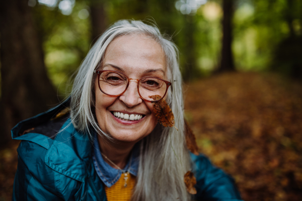 Portrait of happy senior woman with a leaf on her cheek on walk outdoors in forest in autumn.