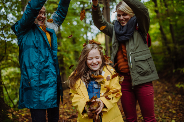 A happy little girl with mother and grandmother having fun with leaves during autumn walk in forest