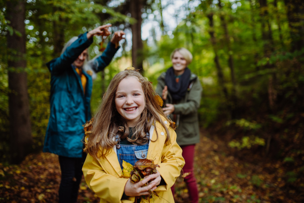 A happy little girl with mother and grandmother having fun with leaves during autumn walk in forest