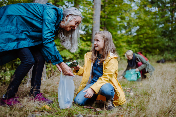 A small girl with mother and grandmother picking up waste outoors in forest.