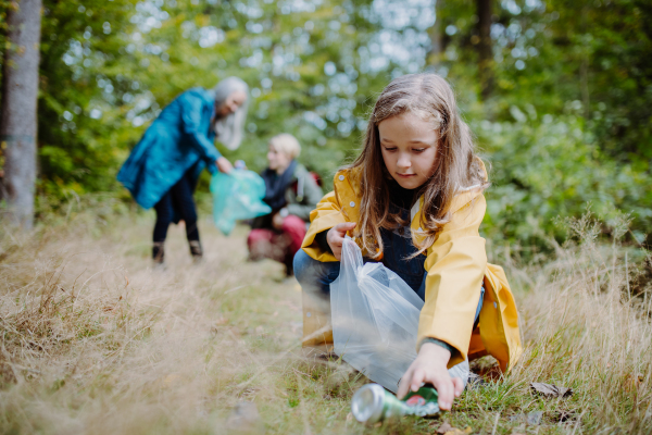 A small girl with mother and grandmother picking up waste outoors in forest.