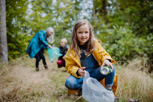 A small girl with mother and grandmother picking up waste outoors in forest.