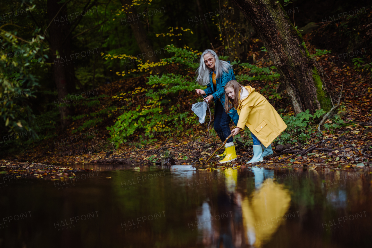 A small girl with grandmother picking up waste from little lake outoors in forest.