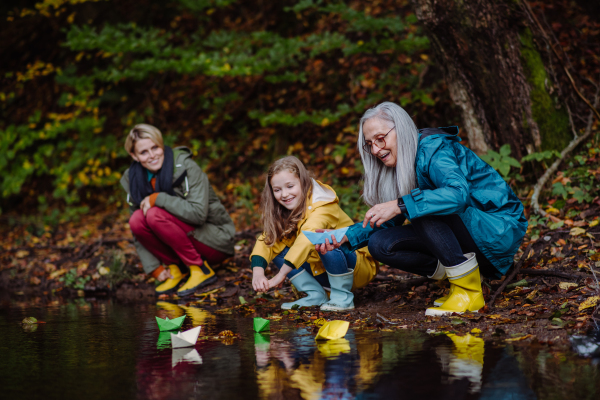 A small girl with mother and grandmother playing with paper boats in lake outoors in nature.