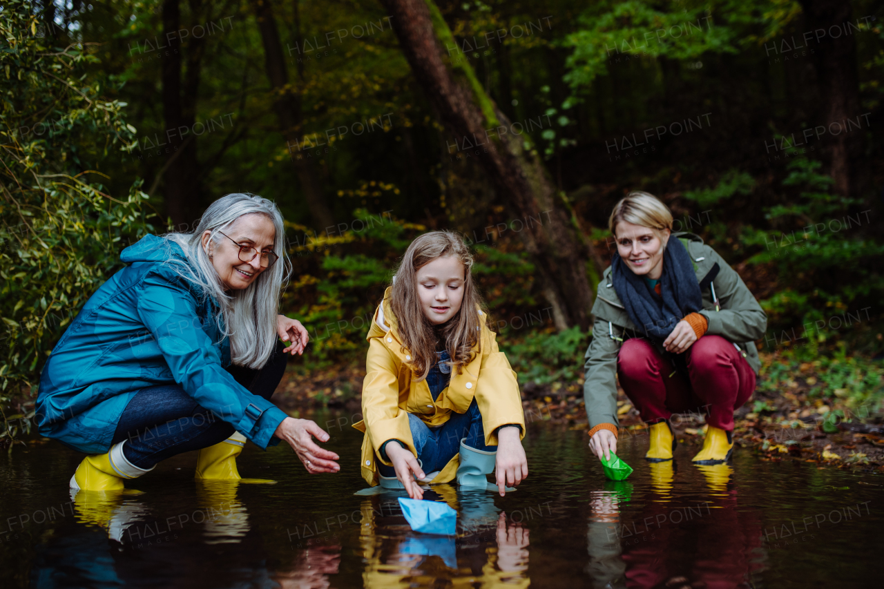 A small girl with mother and grandmother playing with paper boats in lake outoors in nature.