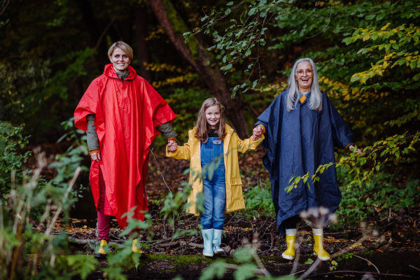 a Portrait of small girl with mother and grandmother in raincoats standing by lake outoors in nature.