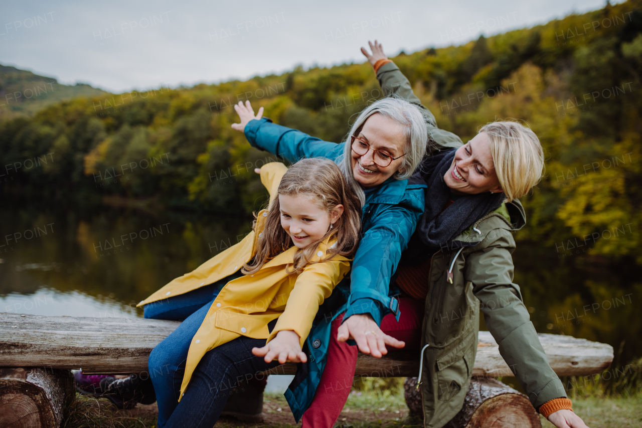 A small girl with mother and grandmother sitting on bench and having fun outoors by lake.