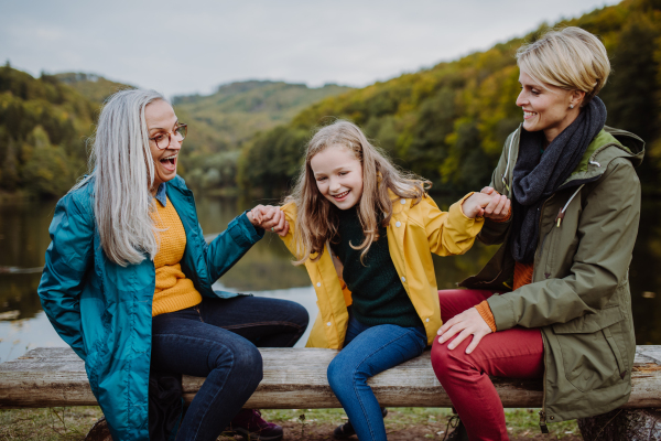 A small girl with mother and grandmother sitting on bench and having fun outoors by lake.