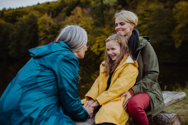 A small girl with mother and grandmother sitting on bench and having fun outoors by lake.