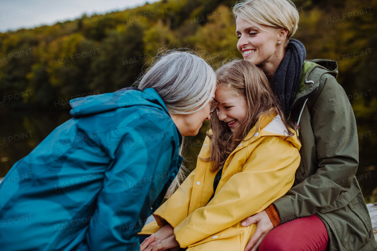 A small girl with mother and grandmother sitting on bench and having fun outoors by lake.