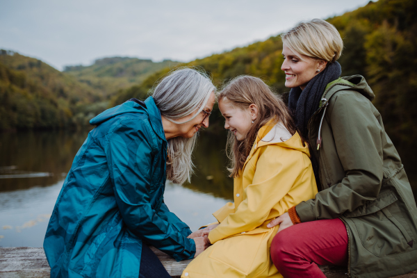 A small girl with mother and grandmother sitting on bench and having fun outoors by lake.