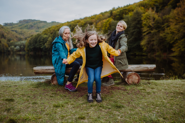 Small girl having fun, her mother and grandmother sitting on a bench outoors by lake.