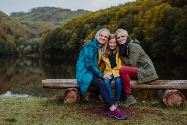 A small girl with mother and grandmother sitting on bench and looking at camera outoors by lake.