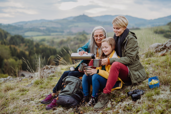 A small girl with mother and grandmother sitting and drinking hot tea on top of mountain in autumn day.