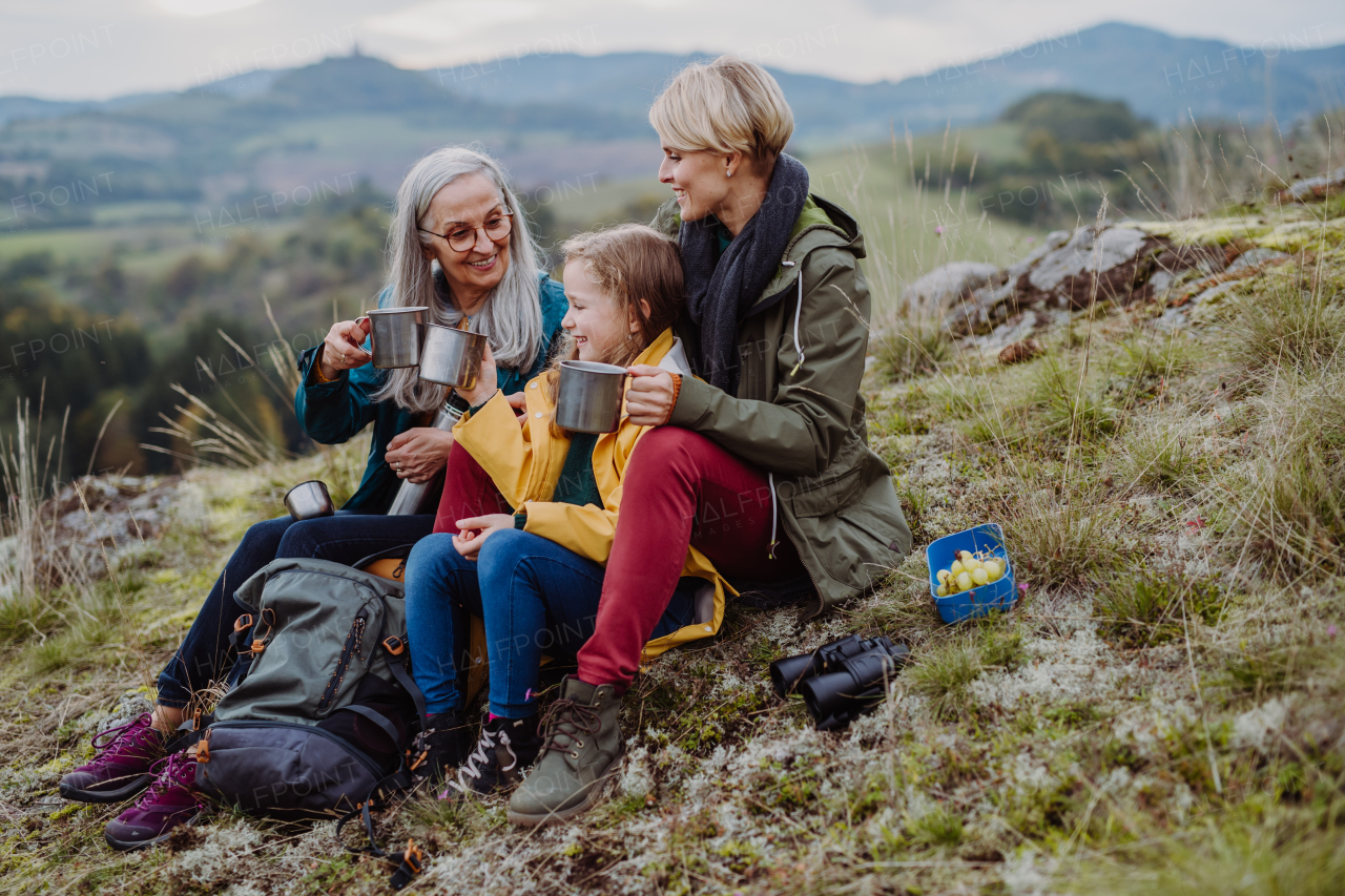 A small girl with mother and grandmother sitting and drinking hot tea on top of mountain in autumn day.