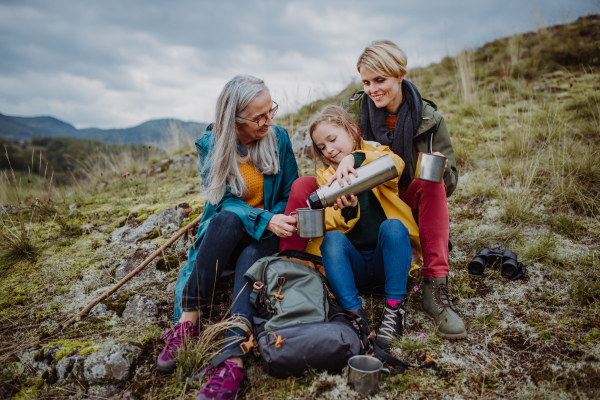 A small girl with mother and grandmother sitting and drinking hot tea on top of mountain in autumn day.