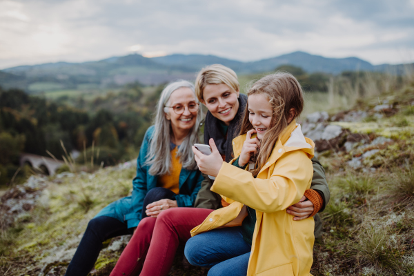 A small girl with mother and grandmother checking smartphone outoors on top of mountain.