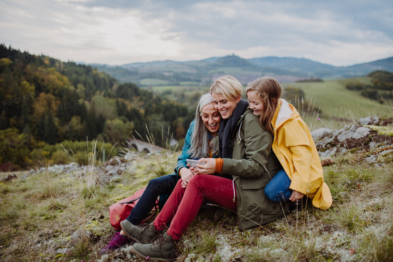 A small girl with mother and grandmother checking smartphone outoors on top of mountain.