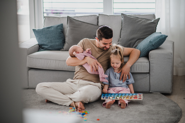 A happy young man taking care of his newborn baby and little daughter indoors at home, paternity leave.