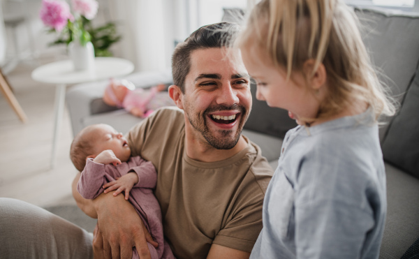 A happy young man taking care of his newborn baby and little daughter indoors at home, paternity leave.