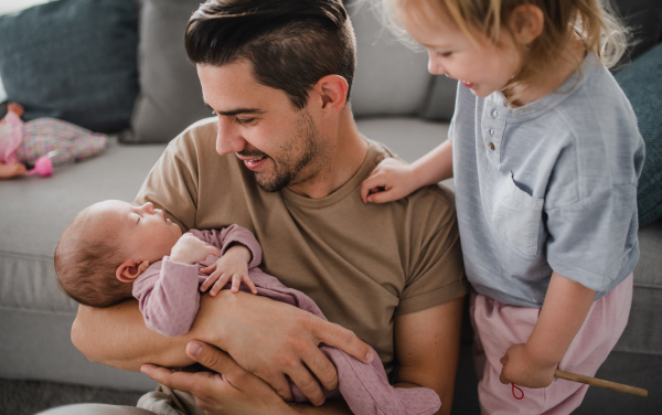 A happy young man taking care of his newborn baby and little daughter indoors at home, paternity leave.