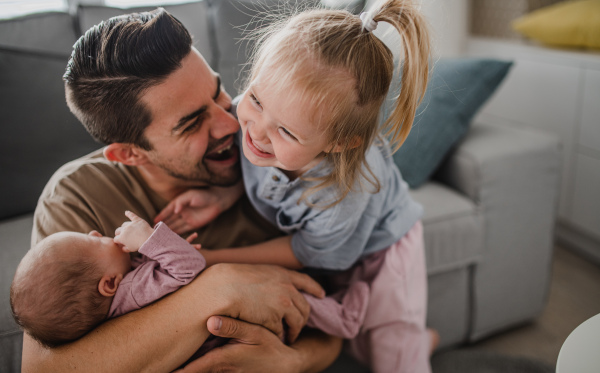 A happy young man taking care of his newborn baby and little daughter indoors at home, paternity leave.