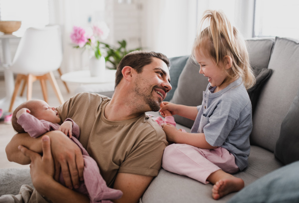 A happy young man taking care of his newborn baby and little daughter indoors at home, paternity leave.