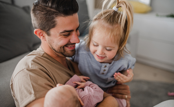 A happy young man taking care of his newborn baby and little daughter indoors at home, paternity leave.