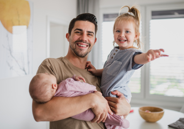 A happy young man taking care of his newborn baby and little daughter indoors at home, paternity leave.