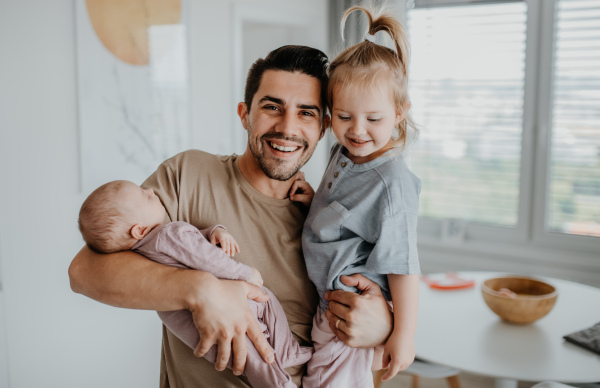 A happy young man taking care of his newborn baby and little daughter indoors at home, paternity leave.