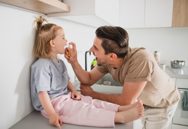 Young father taking care of his little daughter indoors at home, applying a medication.