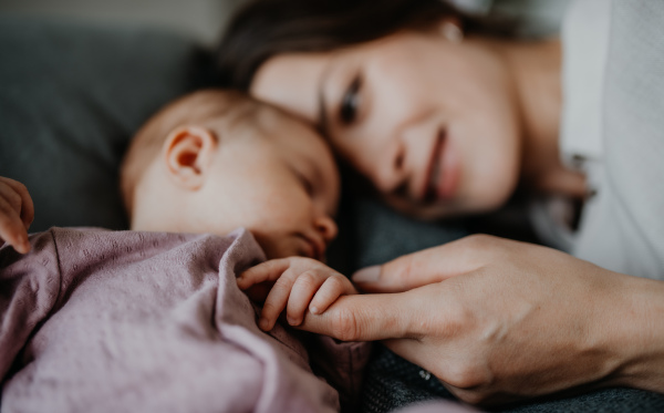 A happy young mother touching her newborn baby girl, lying on sofa indoors at home.