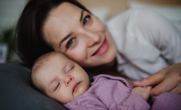 A front view of happy young mother with her newborn baby girl lying on sofa indoors at home, looking at camera.