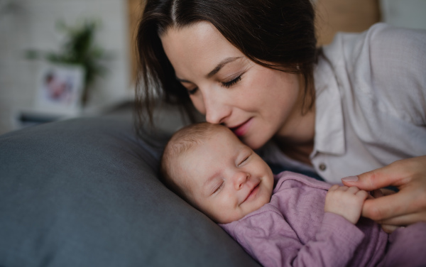 A happy young mother kissing her newborn baby girl, lying on sofa indoors at home.