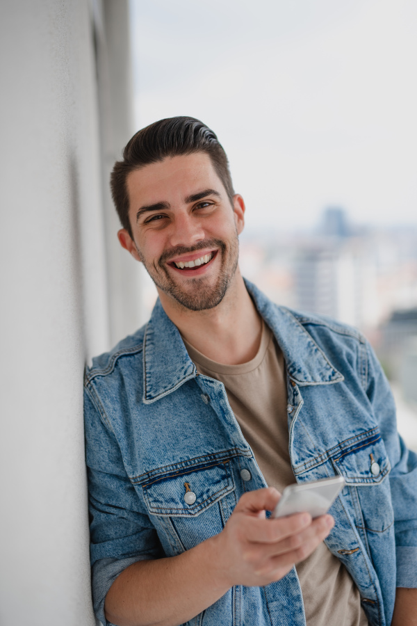 A portrait of young caucasian man with smartphone standing on balcony, looking at camera.