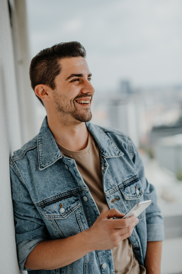 A portrait of happy young caucasian man with smartphone standing on balcony.