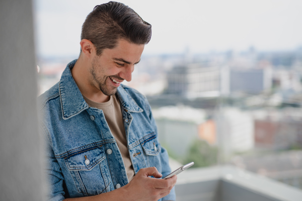 A portrait of young caucasian man with smartphone standing on balcony.