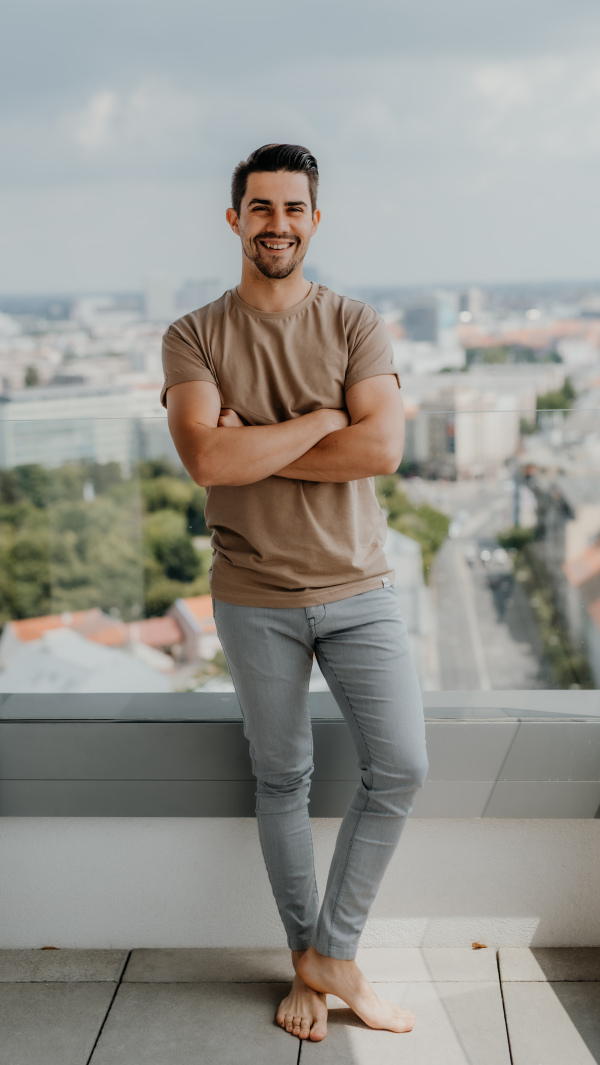 A portrait of happy young caucasian man with arms crossed standing on balcony, looking at camera.