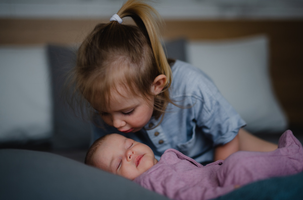 A little girl kissing her newborn baby sister indoors on sofa at home.