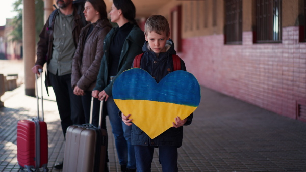 A little refugee boy showing heart shaped Ukrainian flag in station waiting to leave Ukraine due to the Russian invasion of Ukraine