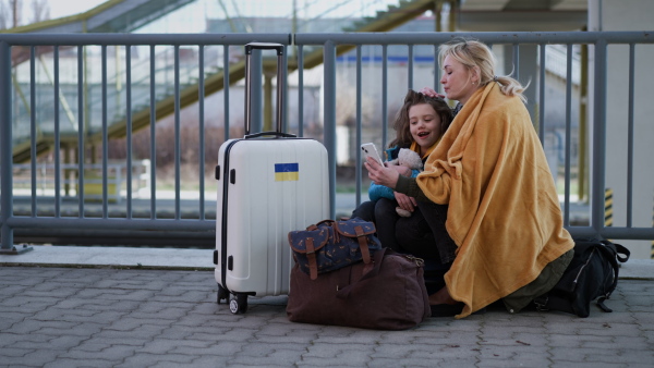 AN Ukrainian immigrants mother with daughter with luggage waiting at train stationand having video call.