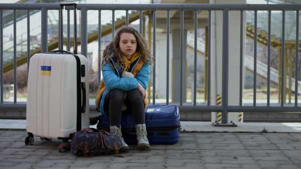 A sad Ukrainian immigrant child with luggage waiting at train station, Ukrainian war concept.