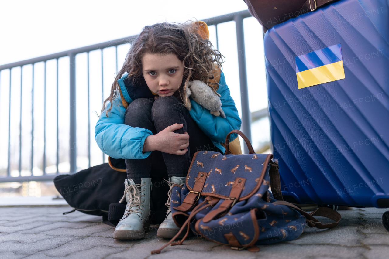 A sad Ukrainian immigrant child with luggage waiting at train station, Ukrainian war concept.