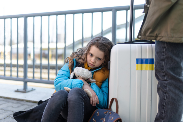 A sad Ukrainian immigrant child with luggage waiting at train station, Ukrainian war concept.