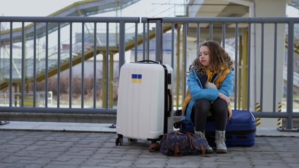 A sad Ukrainian immigrant child with luggage waiting at train station, Ukrainian war concept.