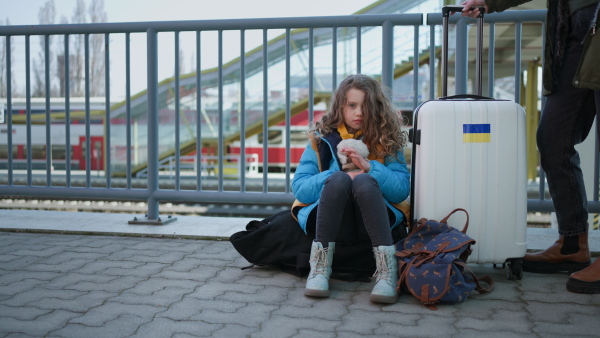 A sad Ukrainian immigrant child with luggage waiting at train station, Ukrainian war concept.