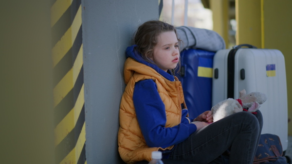 A sad Ukrainian immigrant child with luggage waiting at train station, Ukrainian war concept.