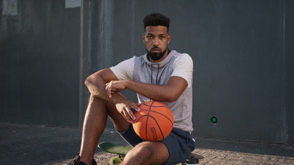 A happy young man with basketball ball outdoors in city, sitting and resting.